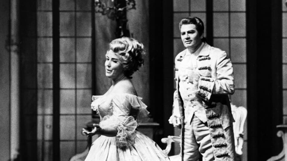 Auf einer Opernbühne: eine Frau und ein Mann sind pompös gekleidet und frisiert. Sie singen.