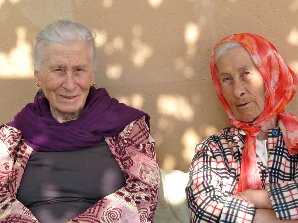 Zwei alte Frauen sitzen, an die Hauswand gelehnt, im Schatten. 