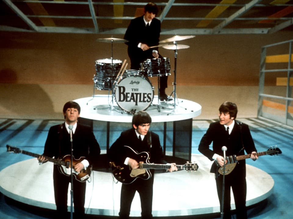 Vier junge Männer im Anzug auf einer Bühne, spielen Gitarre und Schlagzeug.