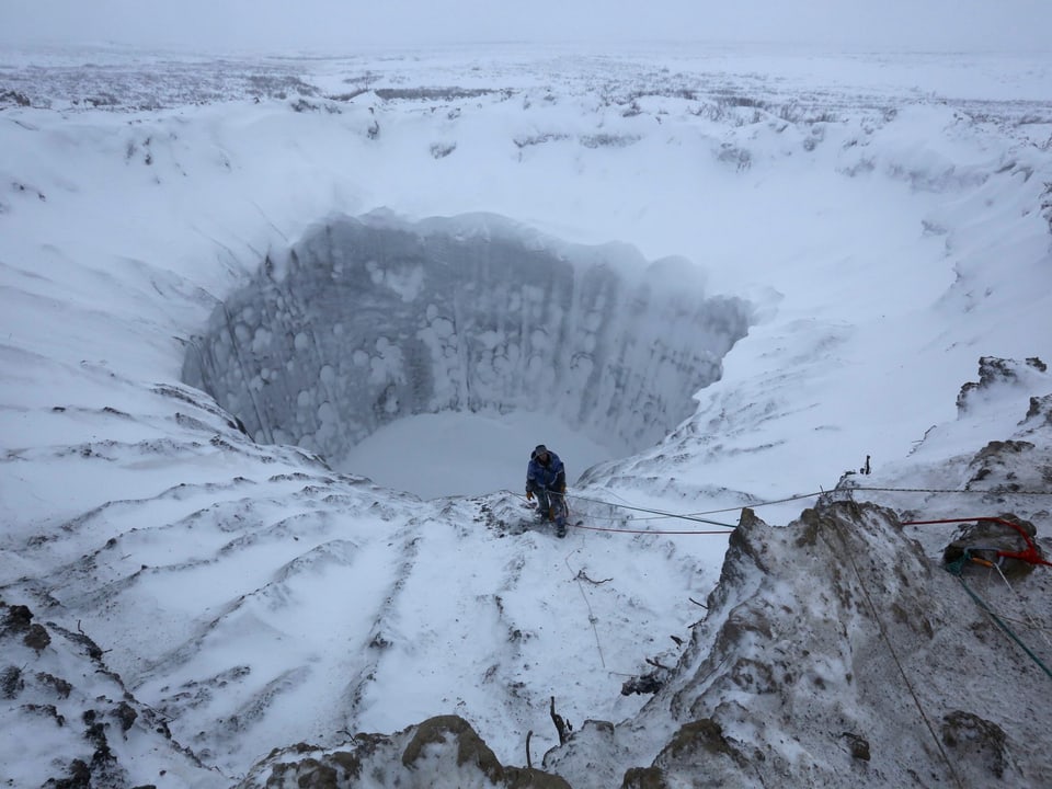 Inmitten einer Schneelandschaft klafft ein grosses, steiles Loch. Am Rand des Kraters steht ein mit Seilen gesicherter Mann. Es scheint, als möchte er sich bald in den Krater abseilen.