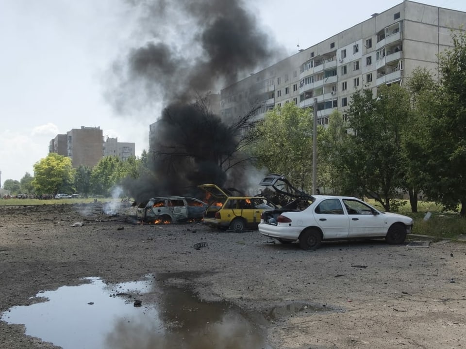 Grosses Wohnhaus, Autos davor brennen nach Bombeneinschlag.