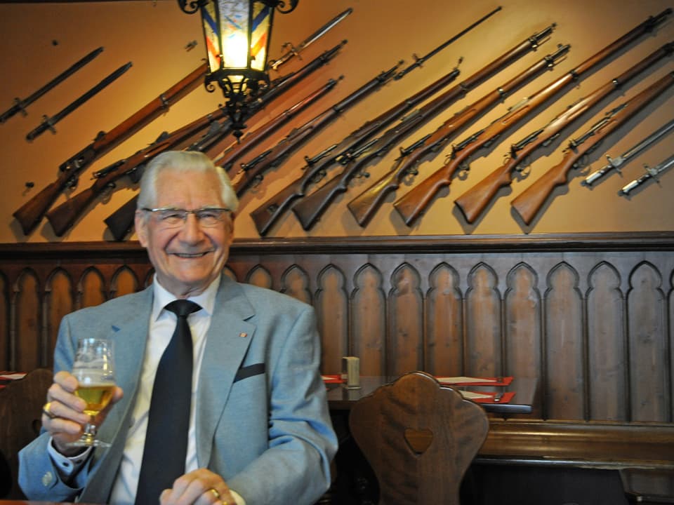 Mann mit Stange Bier sitzt an Holztisch. Gewehre im Hintergrund.