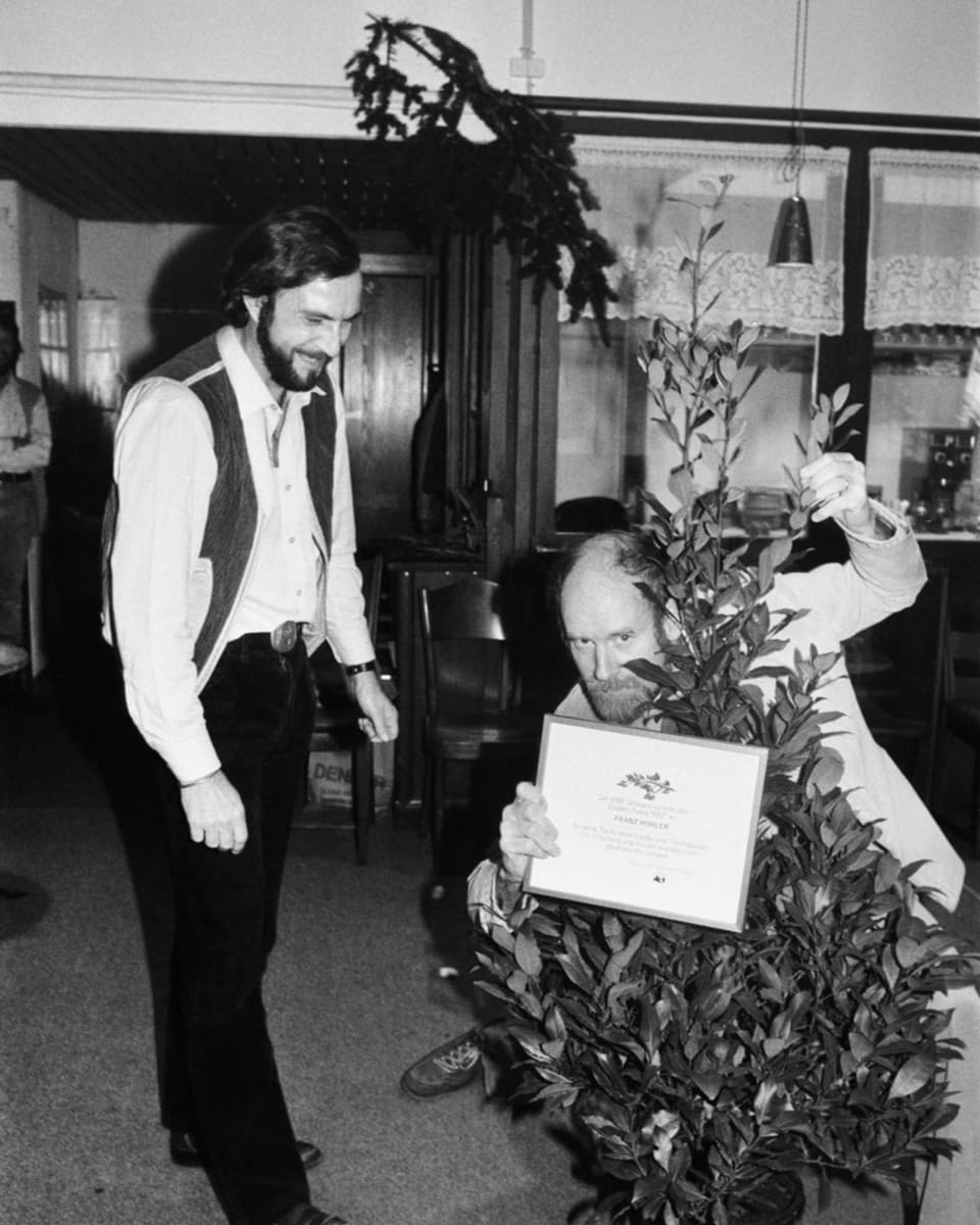 Schwarzweiss-Foto rechts hockt ein Mann mit einem Busch vor sich und Urkunde in der Hand. Links steht ein Mann, lächelnd