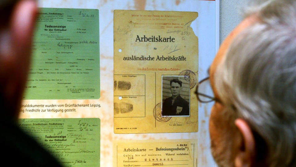 Zwei Männer stehen vor einer Ausstellungswand mit historischen Dokumenten eines Zwangsarbeiters.