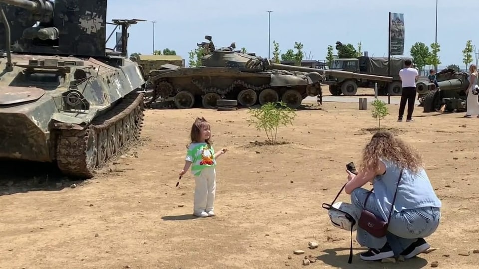 Ein kleines Mädchen steht vor einem Panzer und wird von einer Frau fotografiert.