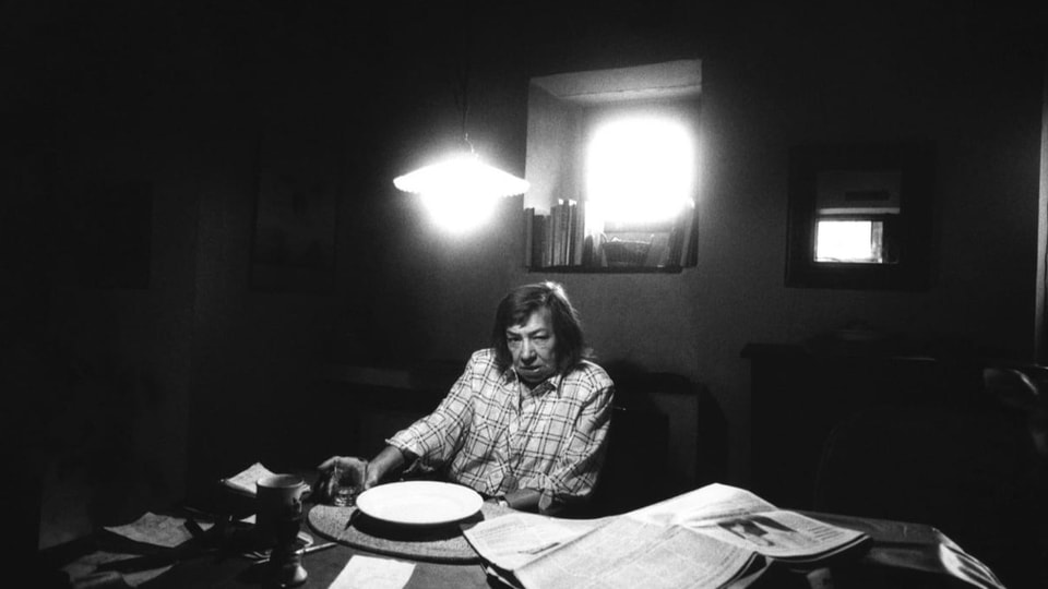 Eine ältere Frau sitzt an einem Schreibtisch unter einer Lampe.