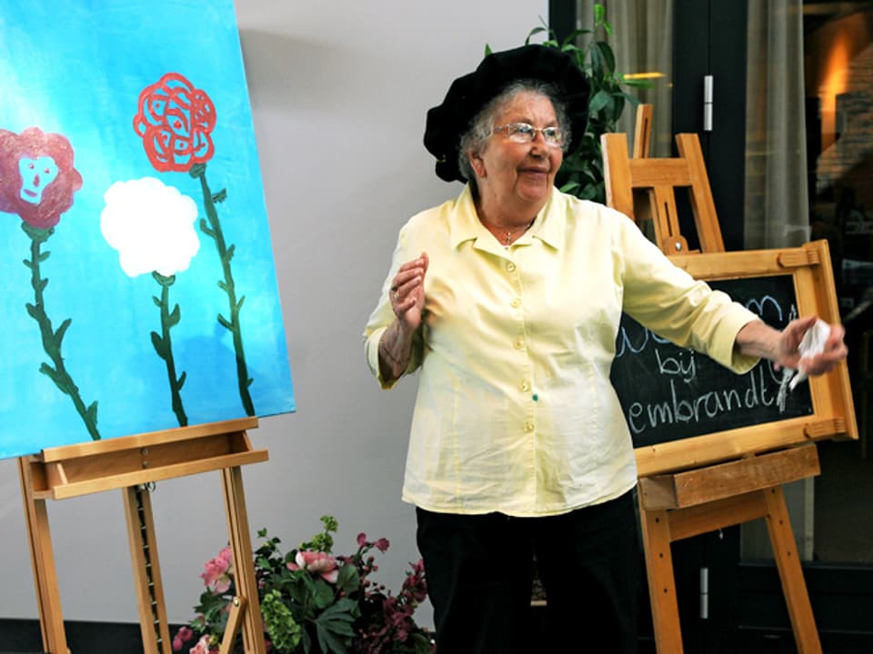 Eine ältere Frau steht vor einer Staffelei, auf der sich ein Gemälde mit drei grossen Blumen befindet.
