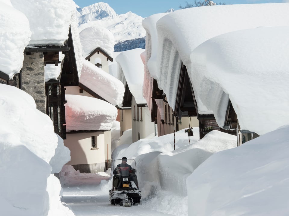 Auf den Dächer in Bedretto türmt sich der Schnee zwei Meter Hoch, ein Snowmobile fährt durch das Dorf.