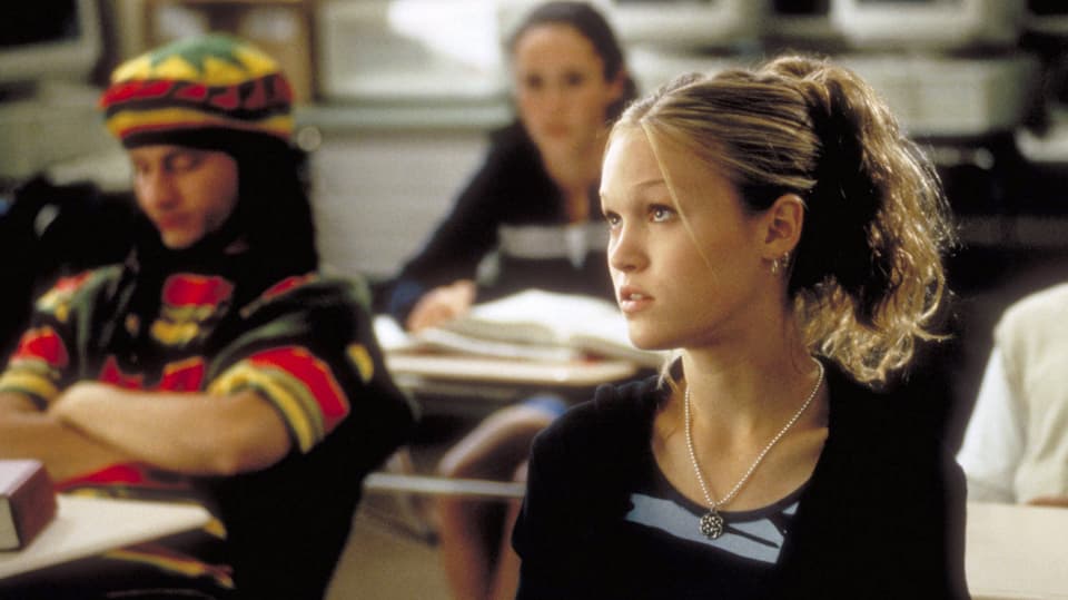 junge Frau links mit blondem, zusammengebundenem Haar, schaut genervt, im Hintergrund Schulbänke.