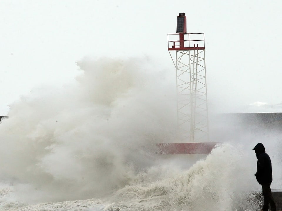 Mann steht an der Küste, daneben prallt eine Welle an Land.
