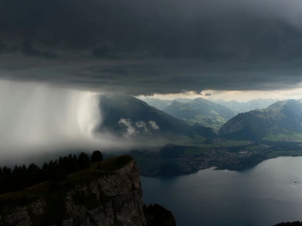 Gewitterwolke mit Niederschlag links über einer Bergregion mit See rechts