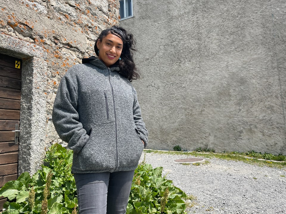 La Nefera freut sich auf das Projekt. La Nefera schaut direkt ins Bild. Sie steht in einem Raum und trägt ein oranges Sweatshirt.