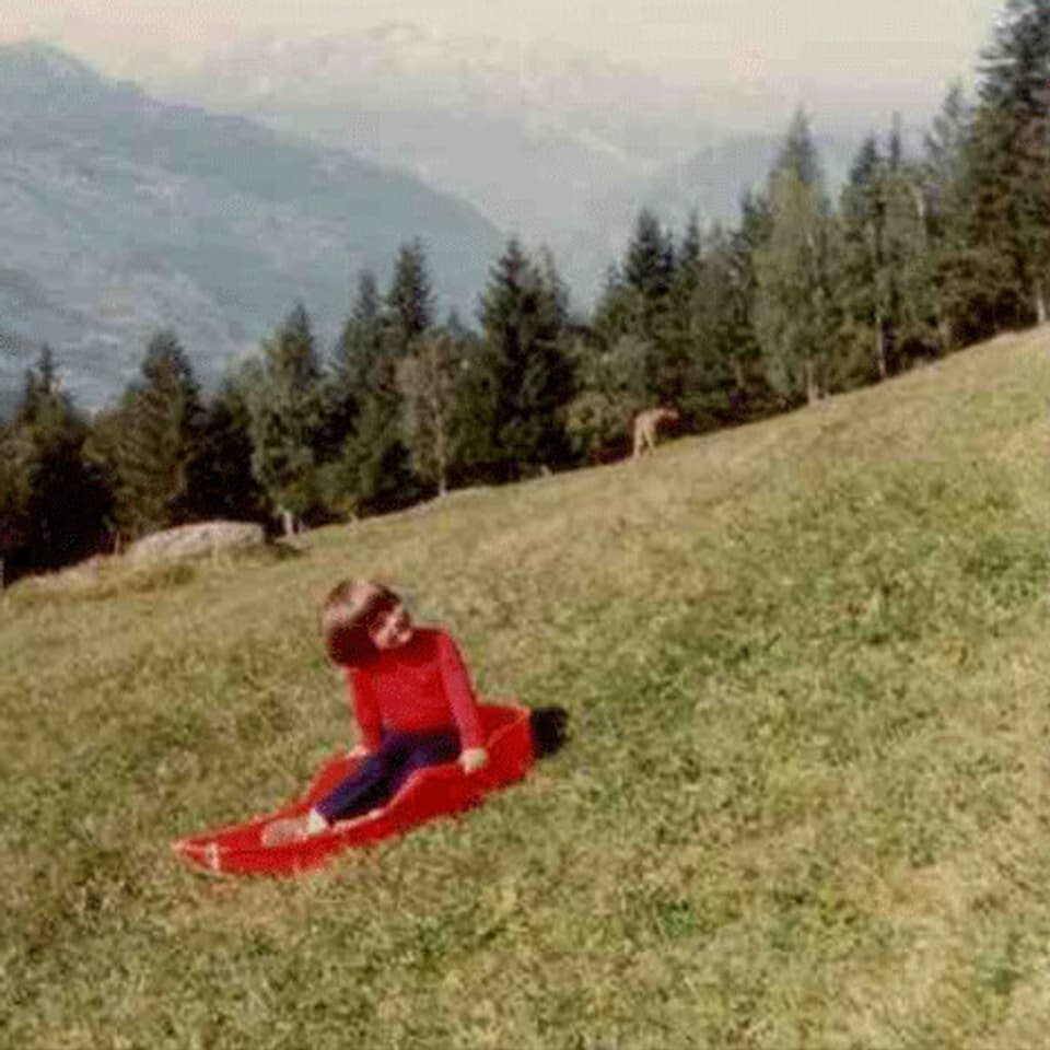 Riccarda als kleines Mädchen sitzt lachend auf einem Bob im Gras.