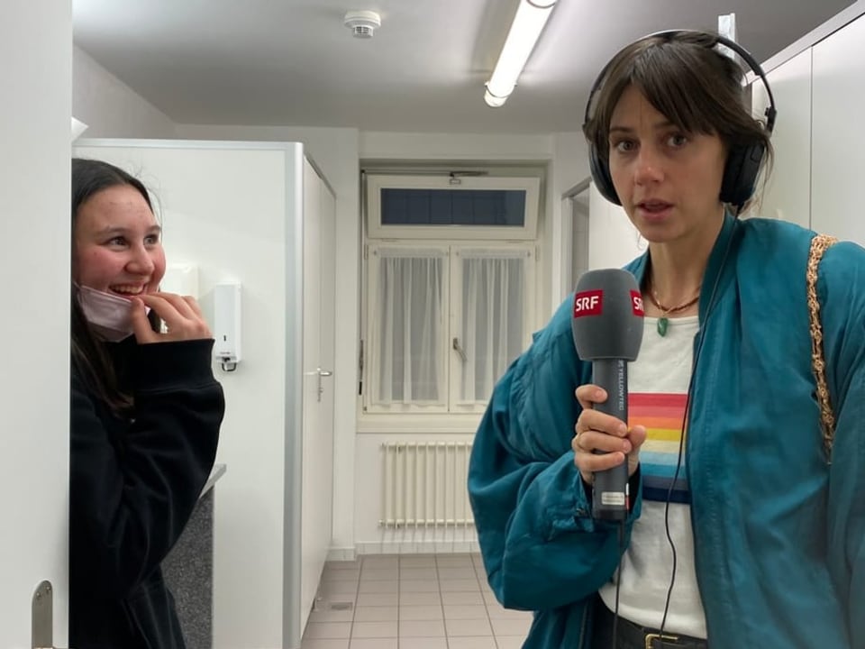 Links steht lachend das schwarz-gekleidete Mädchen. Rechts eine Frau mit Mikrofon in der Hand. Sie stehen in einem Bad.