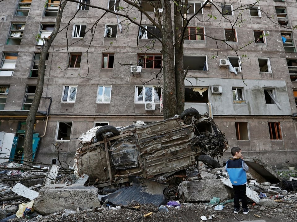 Ein Junge steht neben einem Fahrzeugwrack vor einem beschädigten Wohnhaus in Mariupol.