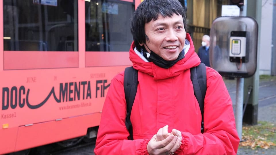 Ein Mann mit roter Jacke steht vor einem roten Tram mit der Aufschrift «documenta fifteen».