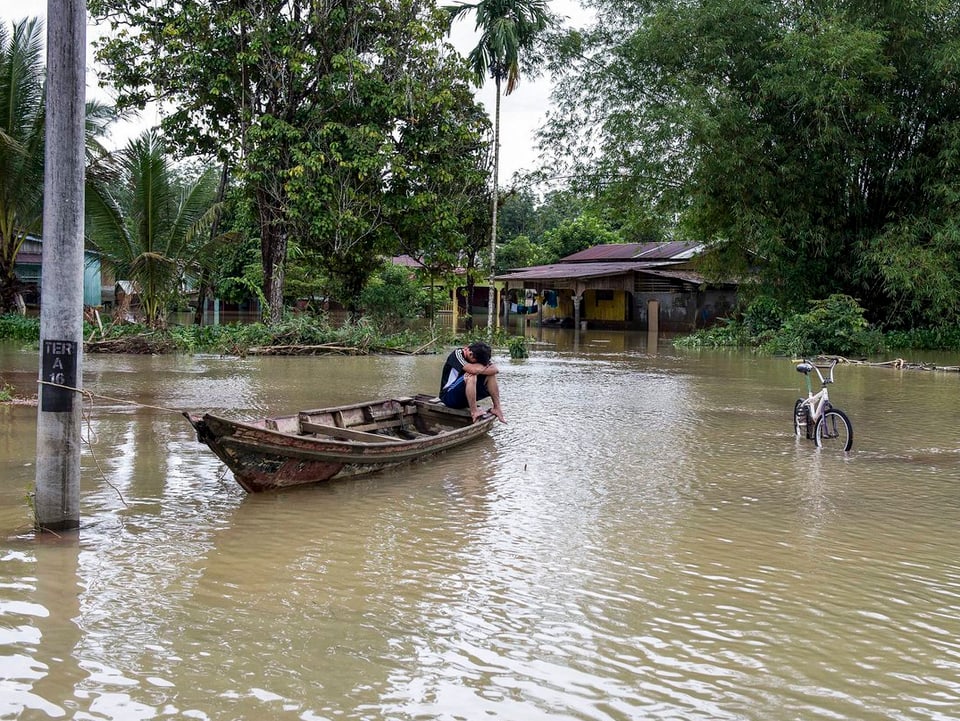 Mann sitzt in einem Holzboot inmitten einer überfluteten Gegend.