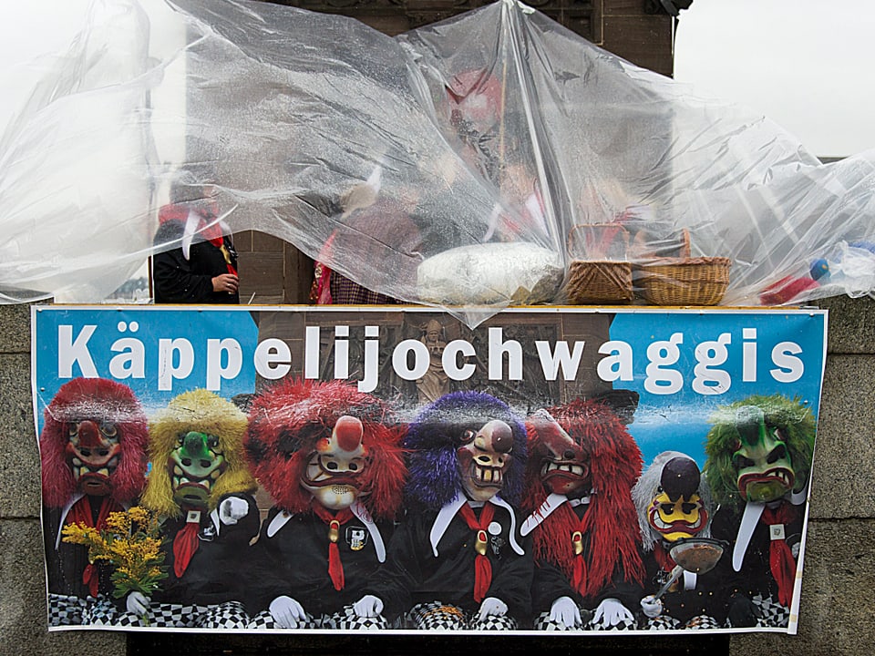 Plakat mit verkleideten Fasnächtlern mit bunten Perücken und Masken. Darüber ist eine Pellerine als Regenschutz gespannt. 