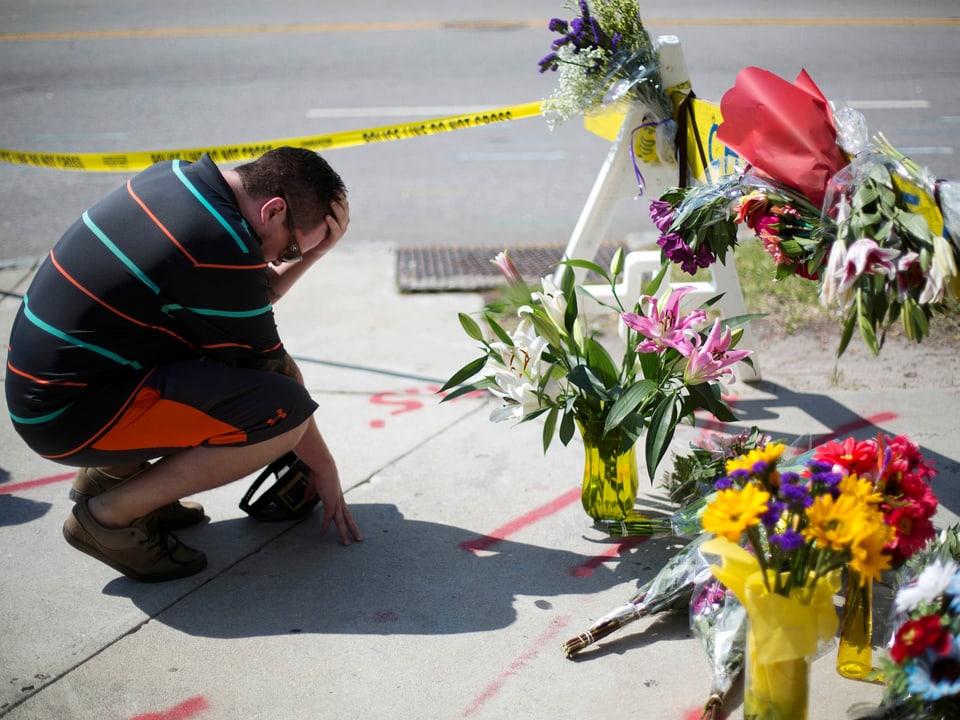 Ein Mann kniet am Boden, vor ihm gelbes Polizeiabsperrband. Auf der Strasse stehen Blumensträusse.