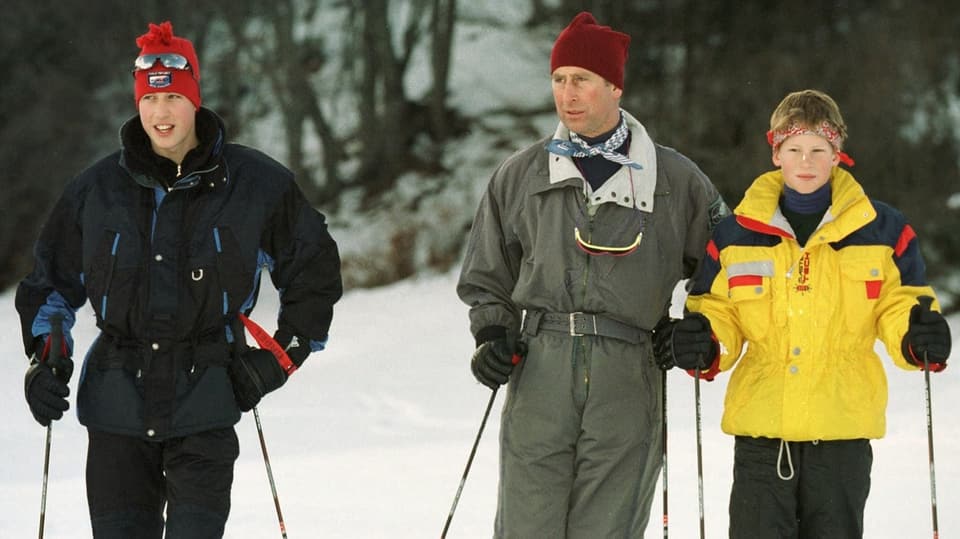 altes Farbfoto, Mitte ein Mann mit grauem Skianzug und roter Mütze, links ein Teenager, rechts ein Junge in gelber Jacke