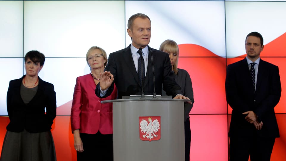 Tusk steht auf einem Podium und spricht in ein Mikrofon, hinter ihm stehen vier seiner Minister.