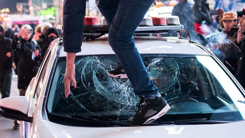 Ein Demonstrant steht in New York auf einem Polizeiauto und bricht die Scheibe mit dem Fuss ein
