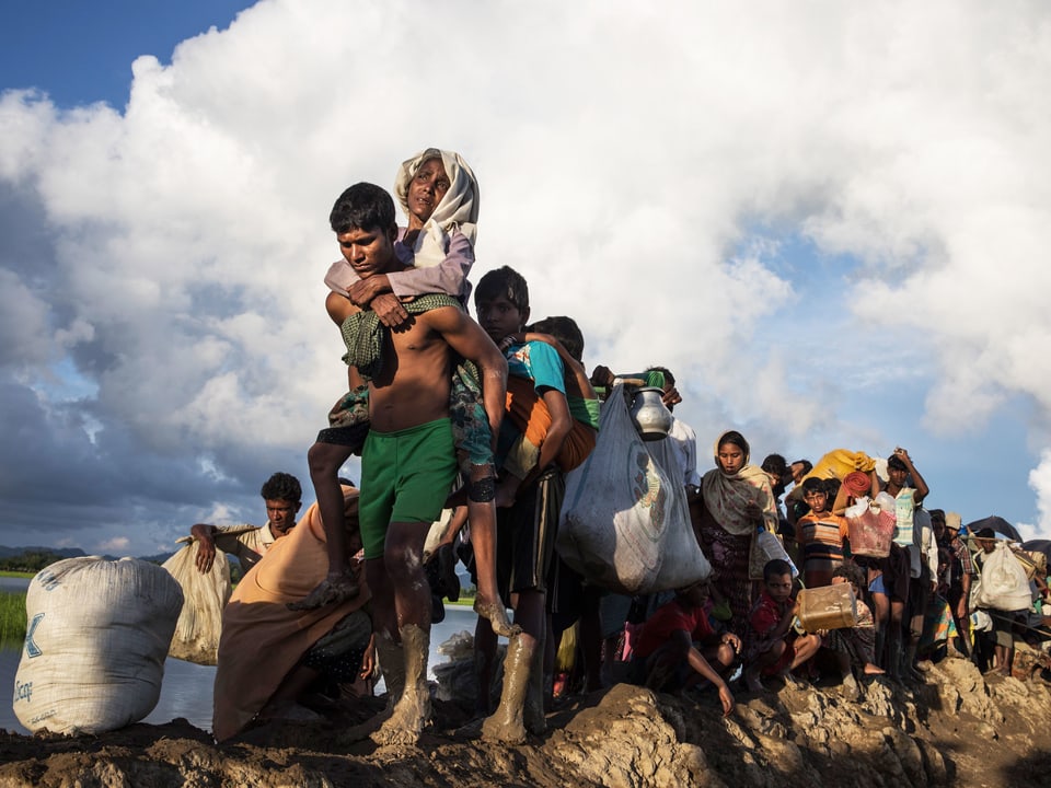 Rohingyas auf der Flucht: Eine Gruppe Menschen geht hintereinander, vorne trägt ein Mann eine alte Frau.