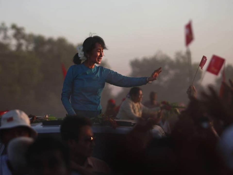 Aung San Suu Kyi sitzt auf einem Auto und winkt in die Menschenmenge. 
