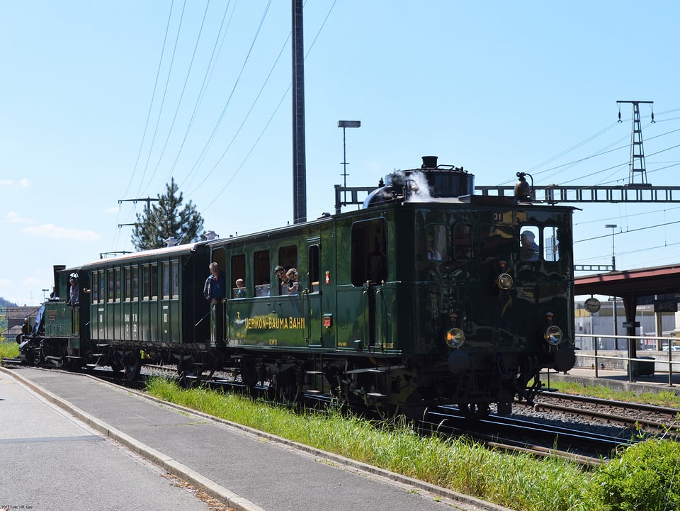 Eine dunkelgrüne elektronische Lokomotive zieht einen einzelnen, alten Waggon.