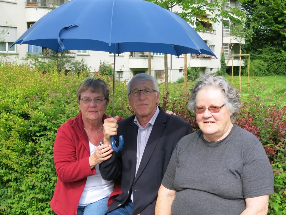 Zwei ältere Frauen und ein älterer Mann mit Sonnenschirm sitzen auf einer Mauer.