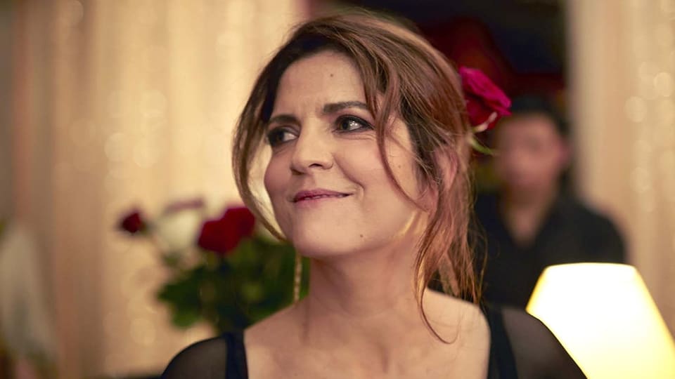Aurore trägt eine rote Rose im Haar und blickt lächelnd an der Kamera vorbei.