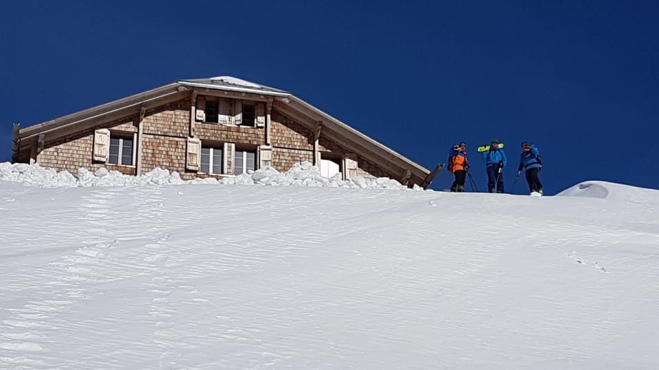 Strahlend blauer Himmel mit einer Hütte im Schnee. Auf der rechten Seite stehen drei Menschen im Schnee.