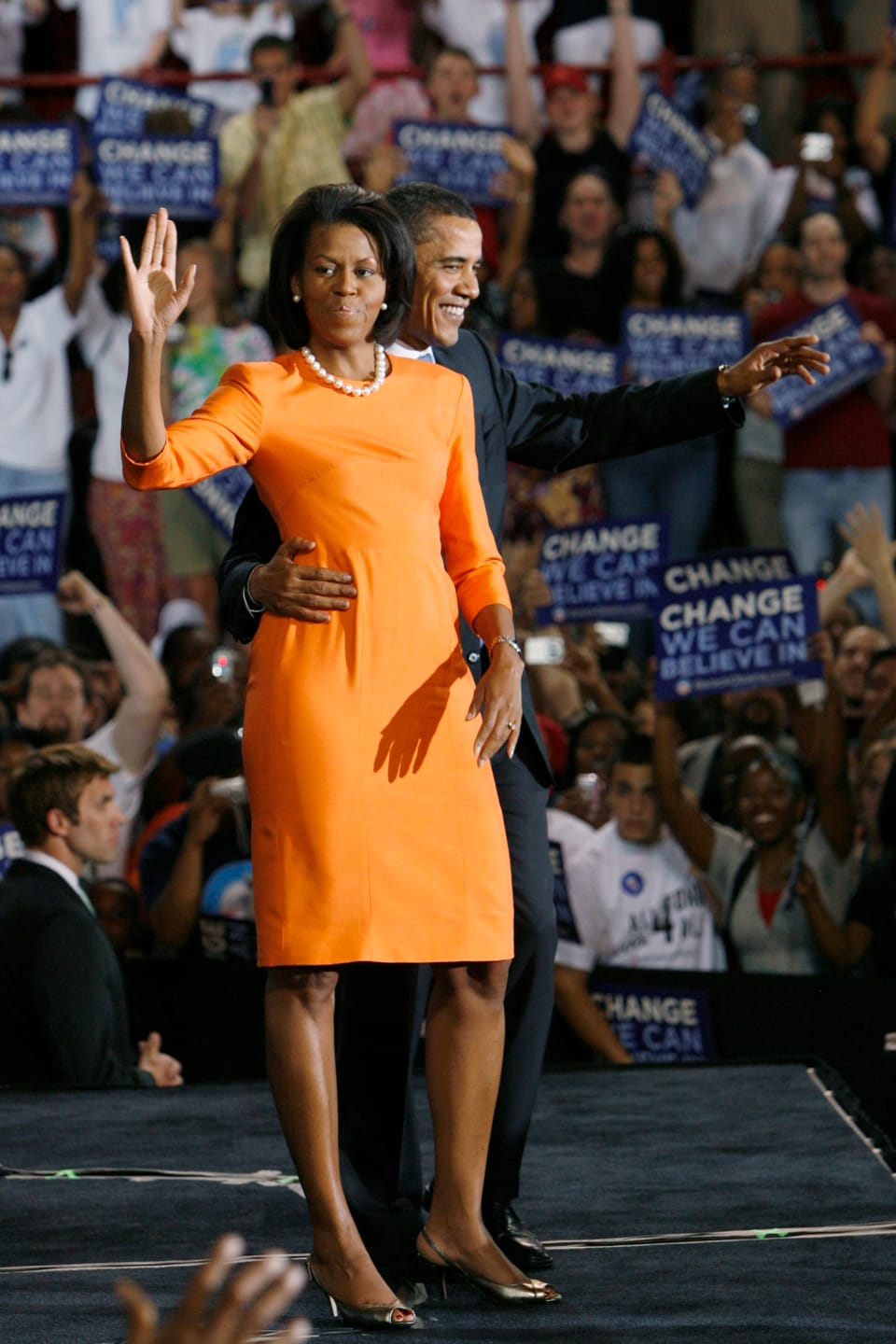 Michelle Obama in einem kurzen, orangenem Kleid winkend. Barrack Obama steht neben ihr und hält sie um die Hüfte.