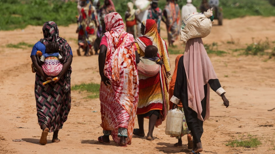  Menschen, hauptsächlich Frauen in farbigen Gewändern, mit Gepäck laufen auf trockenen Boden.