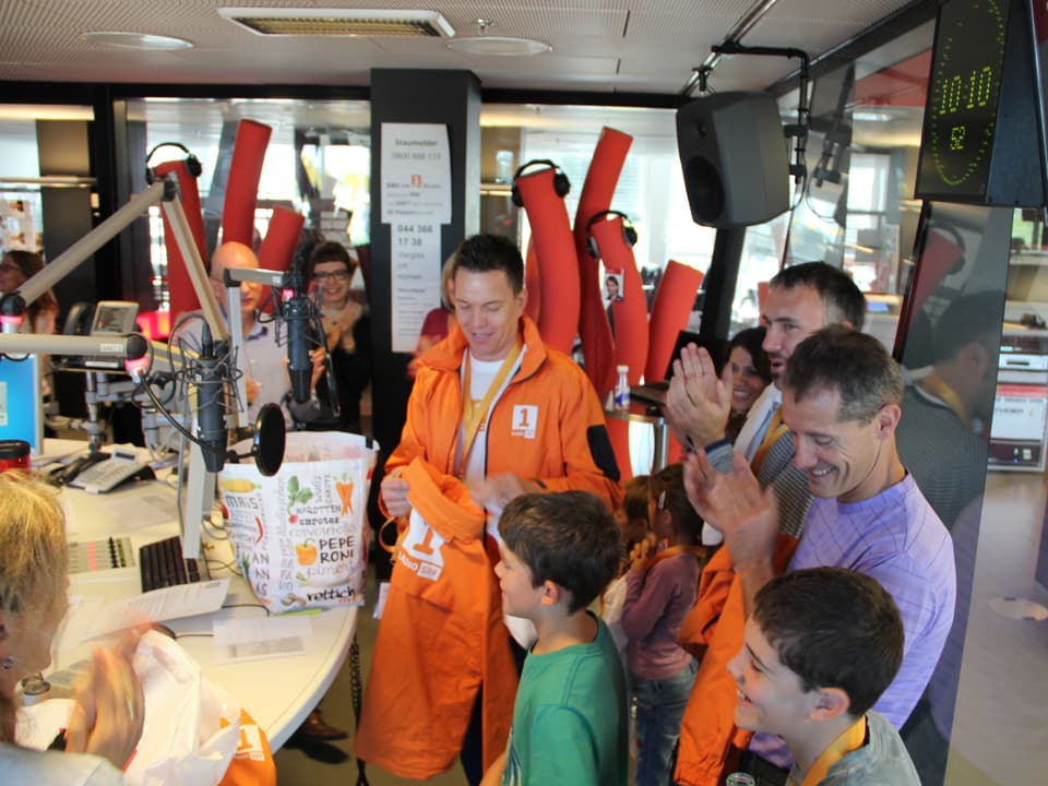 Sven Epiney zieht eine orange Radio SRF 1-Jacke aus der Einkaufstasche.