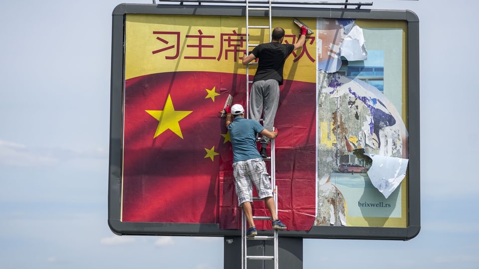 Zwei Personen installieren ein Werbeplakat mit chinesischer Flagge und Schriftzug auf einem Werbetafel.
