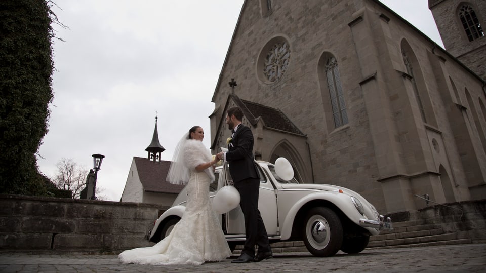 Iva Bencic im weissen Hochzeitskleid und Sandro Cavegn stehen vor einem weissen VW Käfer