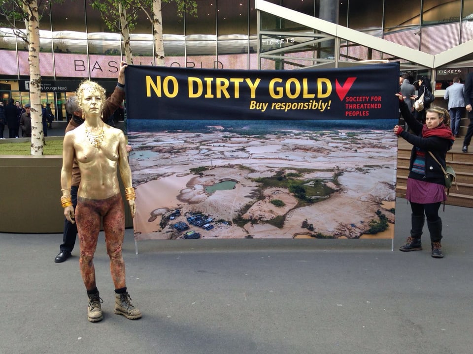 Frau mit goldener Bodypaintingfarbe am Körper steht vor einem Plakat mit der Aufschrift "no dirty gold"