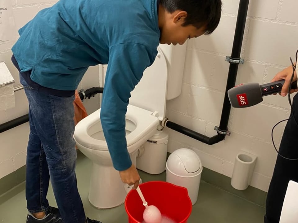 Der Junge steht vor einem WC. In der Hand hält er die WC Bürste, die in einem roten Kübel voll Seife liegt.