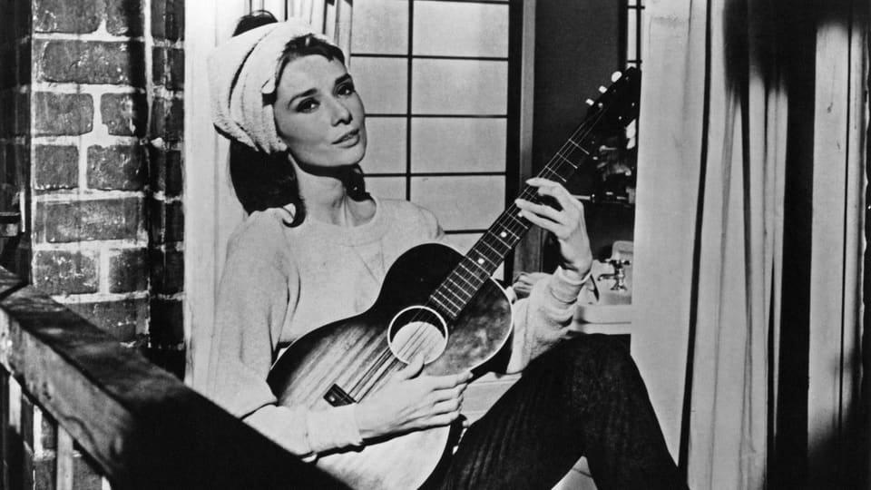 Schauspielerin Audrey Hepburn sitzt auf einem Fenstersims und spielt Gitarre, dabei blickt sie nachdenklich in die Ferne