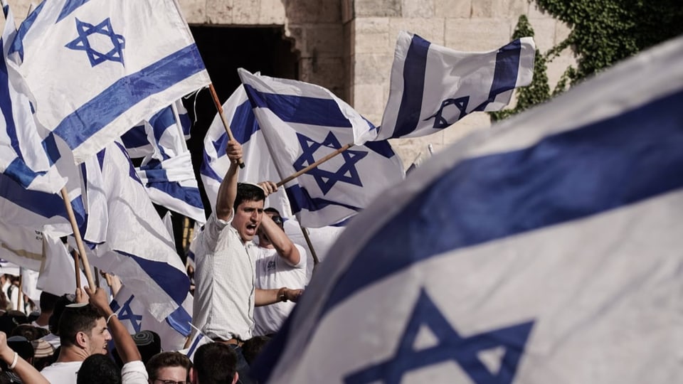 Menschenmenge mit weiss-blauen Flaggen mit Stern (Israel), manche rufen etwas, vor heller Steinmauer.
