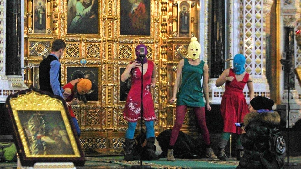 Vier Frauen in bunten Sturmhauben stehen vor einem goldenen Altar. Sie singen und tanzen. Ein Wachmann greift ein.