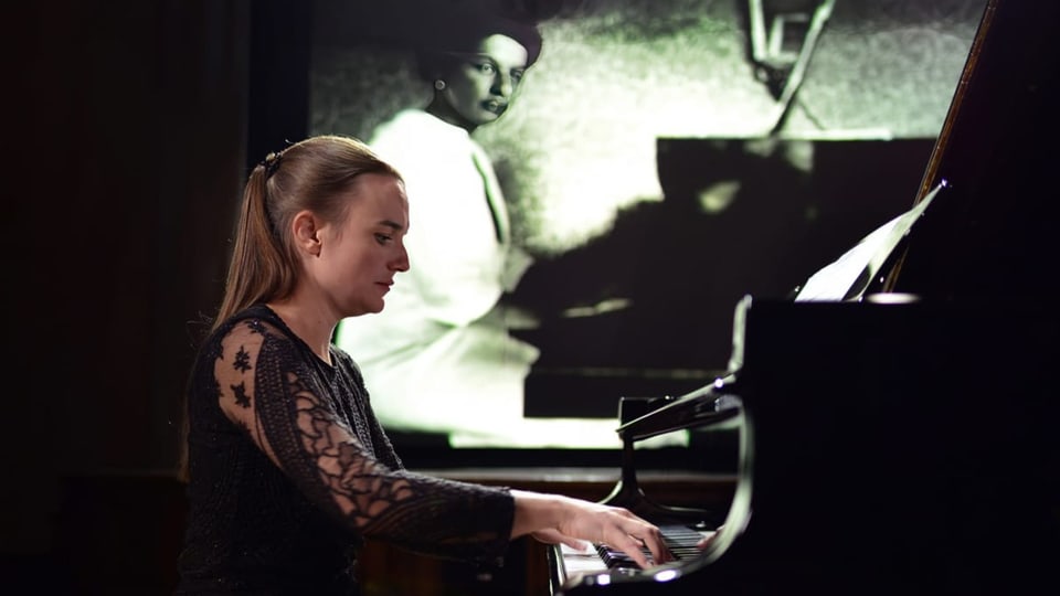 Eine Pianistin spielt an schwarzem Flügel; im Hintergrund sieht man ein Foto einer Frau, die ebenfalls am Flügel sitzt.