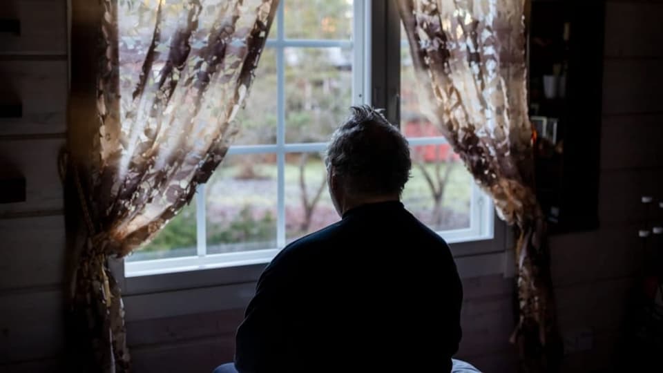 Ein alter Mensch schaut in einem abgedunkelten Raum aus dem Fenster.