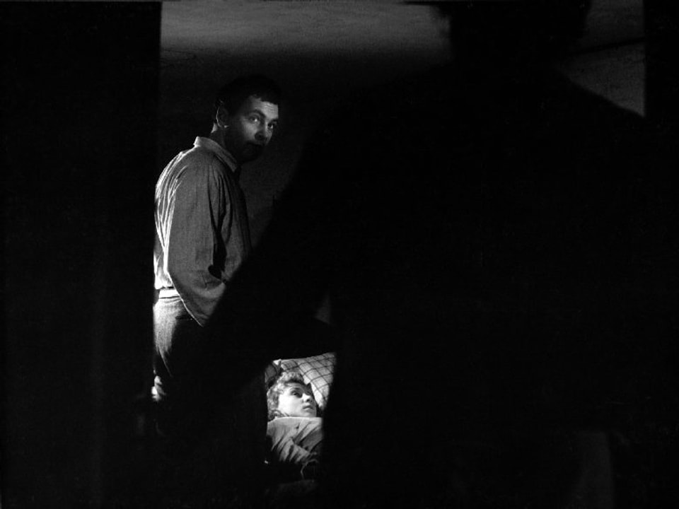 Ein Mann steht neben einem Bett, in welchem eine Frau liegt. Im Vordergrund verdeckt eine Silhouette den Grossteil des Bildes.