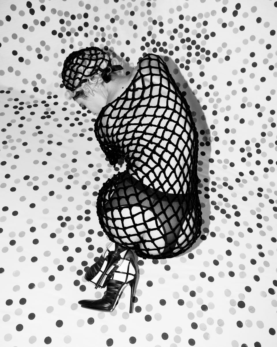 Schwarzweissbild: Eine Frau in Netzkleid kniet auf einem gepunkteten Boden