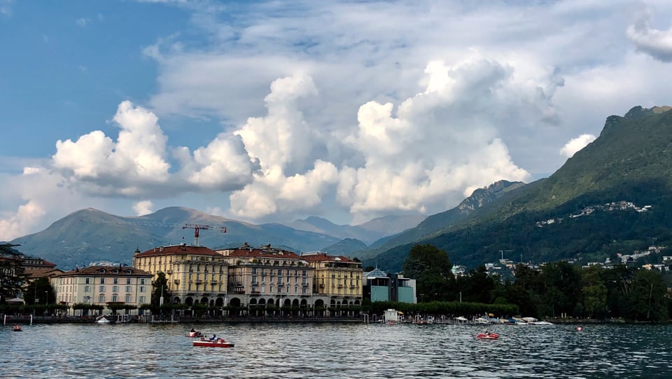 Blick über den Luganersee zur Stadt Lugano, dahinter grosse Quellwolken.