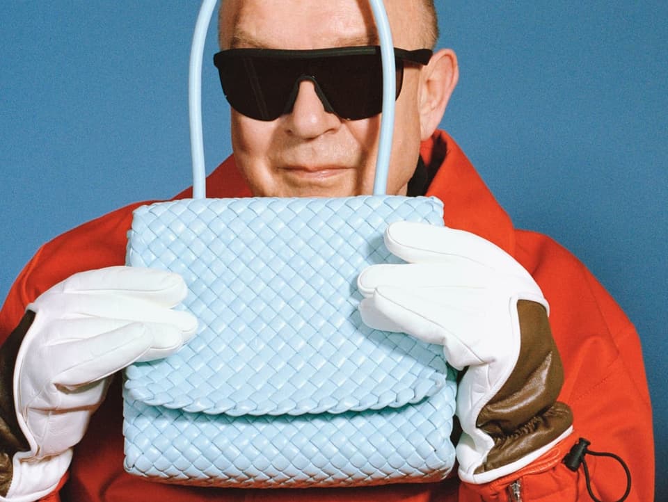 Älterer Herr mit Sonnenbrille, roter Jacke und Handschuhen versteckt sich hinter Handtasche