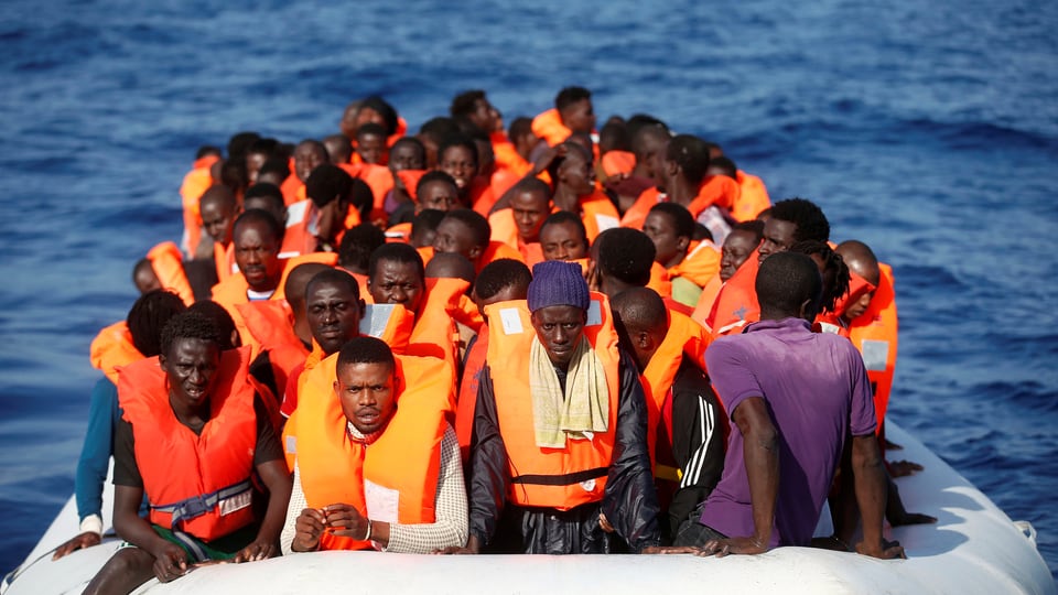 Eine Gruppe Schwarzafrikaner sitzt im Schlauchboot. Sie trägt orange Sicherheitswesten.