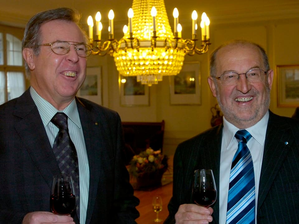 Zwei Männer mit Glas Wein, im Hintegrund Kronleuchter.